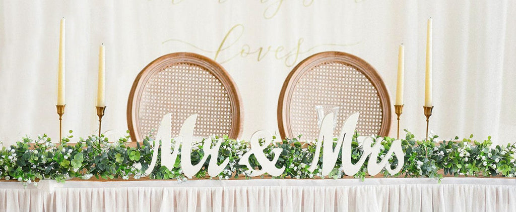 Faux eucalyptus garland with white flowers and 'Mr & Mrs' wooden letters on a table with candles and chairs.