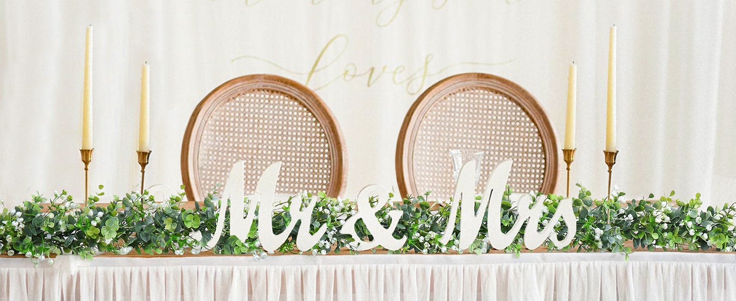 Faux eucalyptus garland with white flowers and 'Mr & Mrs' wooden letters on a table with candles and chairs.