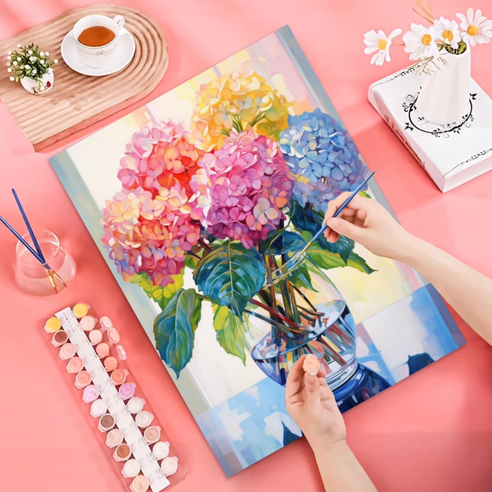 Paint-by-number kit with colorful hydrangeas in a vase, hands painting, and paint pots on a pink background.