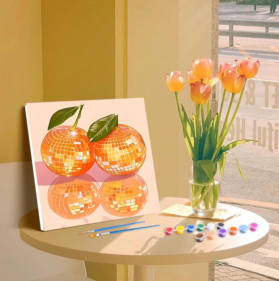 Paint by number kit with two disco ball oranges on canvas, brushes, and acrylic paints on a table with tulips.