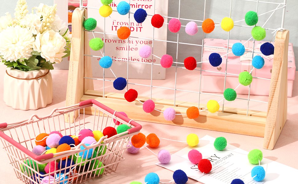 Rainbow pom pom garlands displayed on a wooden grid and in a pink basket, with a floral arrangement to the left.