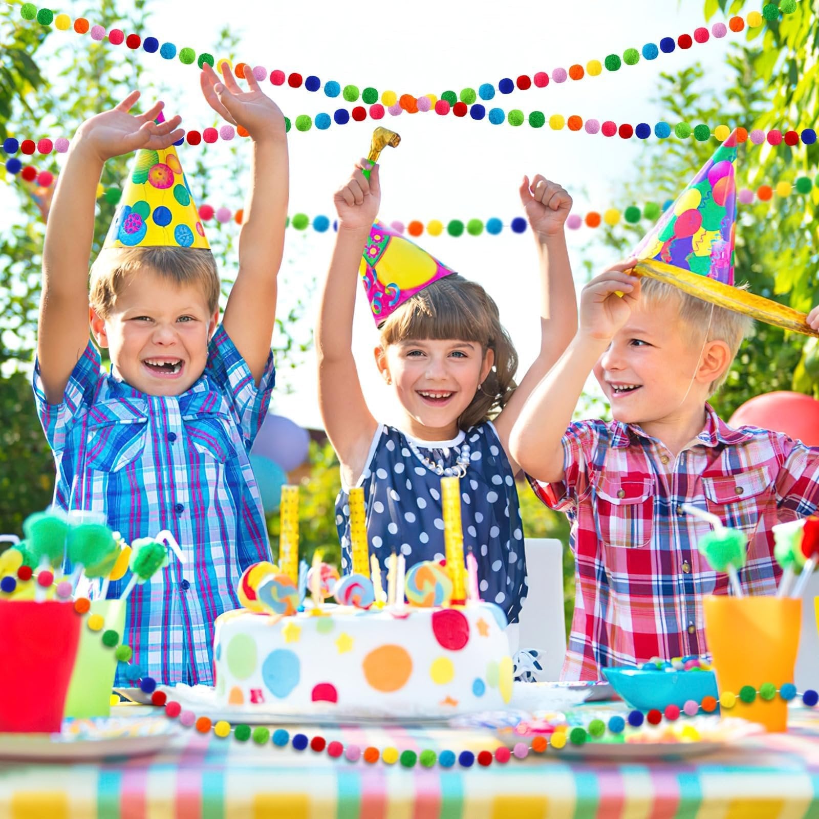Three children in party hats celebrating around a cake outdoors, with colorful pom pom garlands decorating the scene.