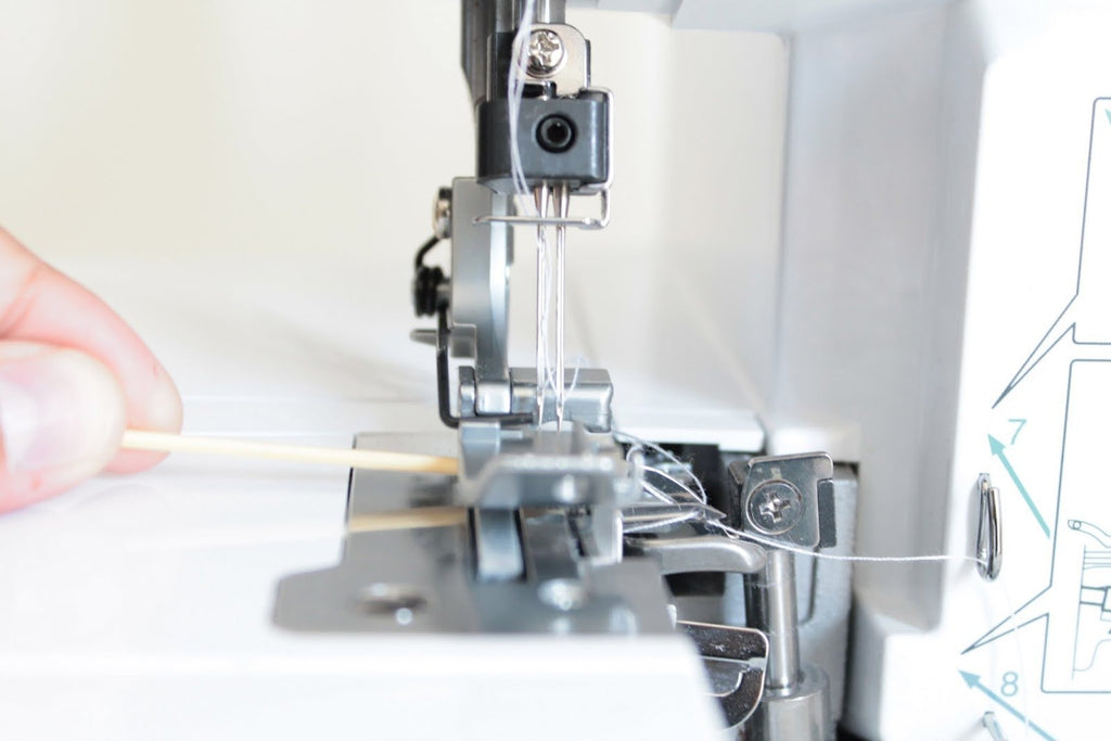 Close-up of a hand using a wooden stick to guide white thread through a sewing machine needle assembly.