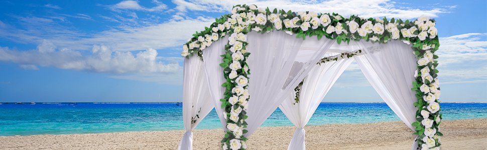 White artificial rose vine garlands draped on a wedding arch on a beach with blue ocean and sky.