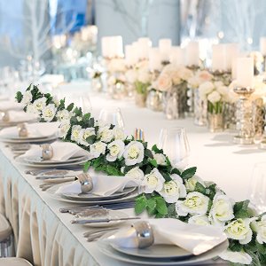 White artificial rose vine garlands with green leaves draped across a formal table setting with plates, silverware, and candles.
