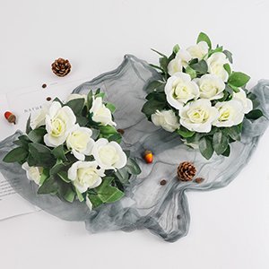 Two artificial white rose vine garlands with green leaves arranged on a grey cloth, with pinecones and acorns.