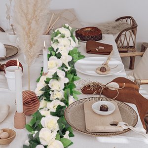 White artificial rose vine garland with green leaves draped across a decorated table setting with candles and plates.