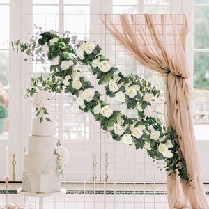 Wedding cake and white rose garlands draped on a grid arch with beige fabric.