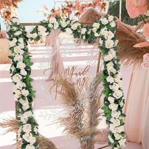 White artificial rose vine garlands with green leaves decorating a wedding arch with pampas grass and pink flowers.
