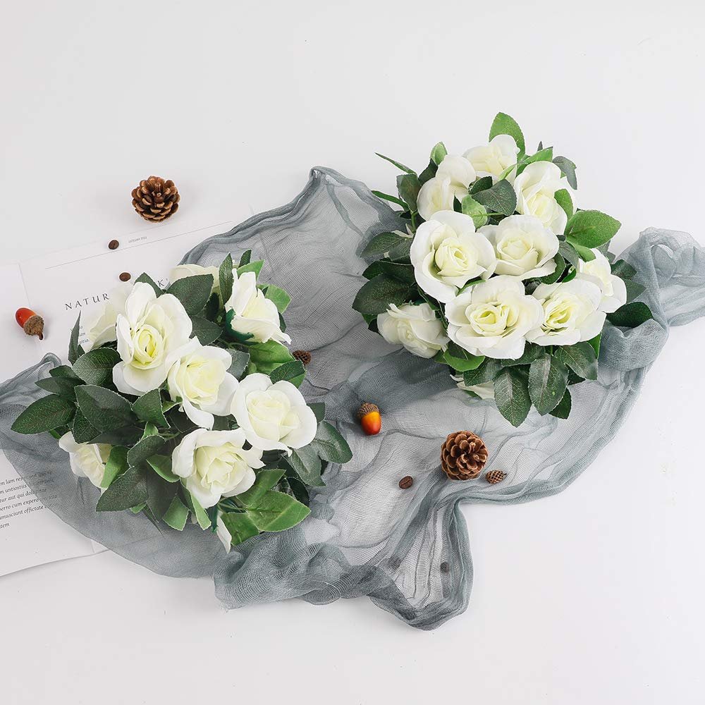 Two bouquets of white artificial roses with green leaves arranged on a grey sheer cloth with decorative acorns and pinecones.