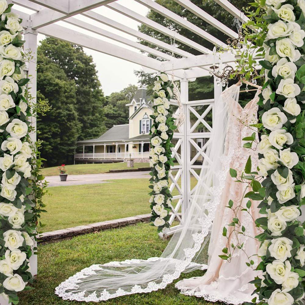 White artificial rose garlands draped on a white pergola with a wedding dress and veil hanging nearby.