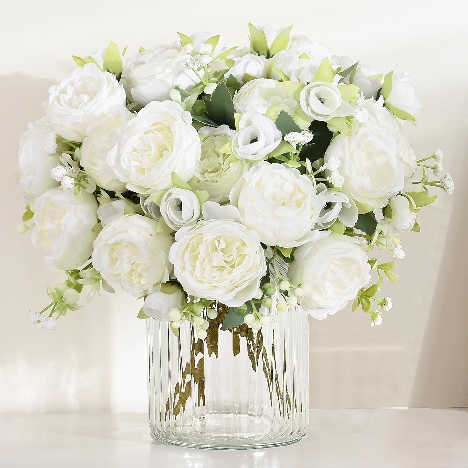 White silk peonies bouquet in a clear ribbed glass vase on a white surface, with soft lighting.
