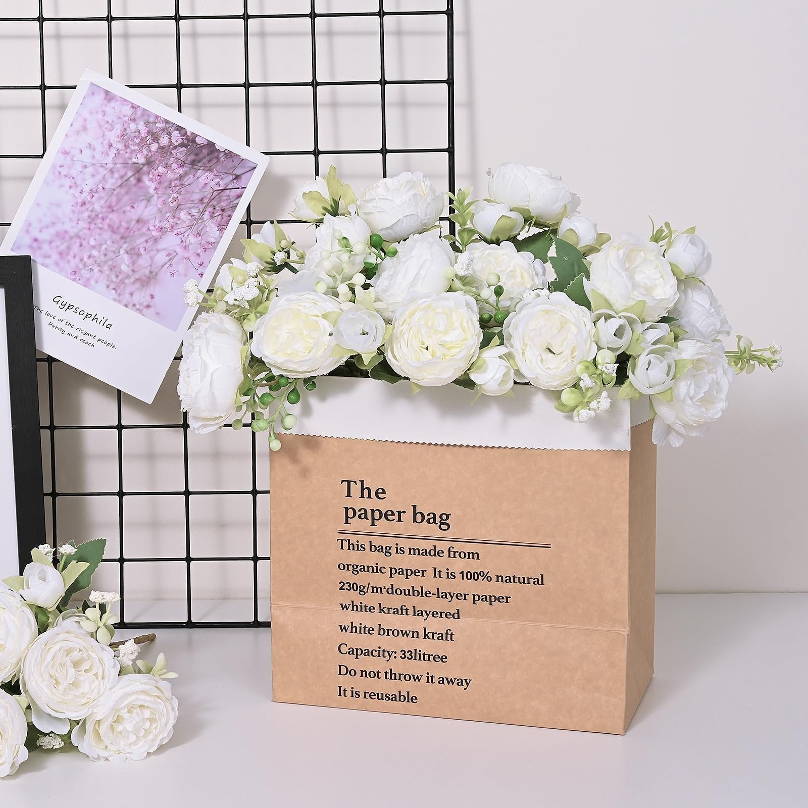 White artificial peony bouquet arranged in a brown paper bag planter, with a decorative card behind it.