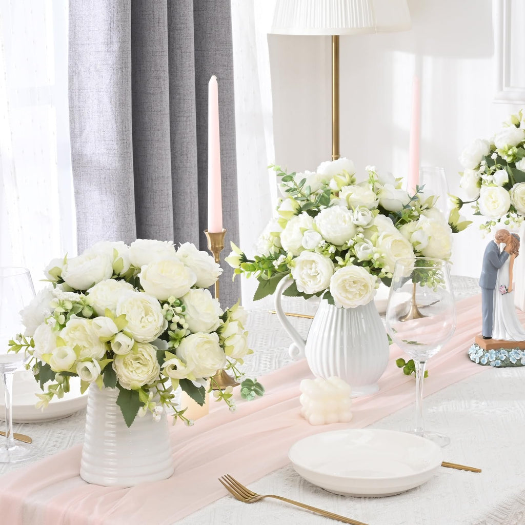 Two white artificial peony bouquets in white vases on a table setting with pink runner, candles, and a wedding cake topper.