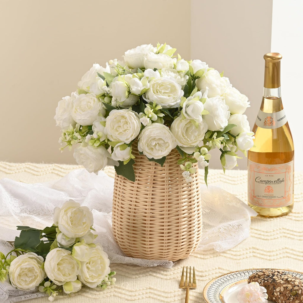 White artificial peony bouquet in a woven basket with a bottle of wine and a plate of bread