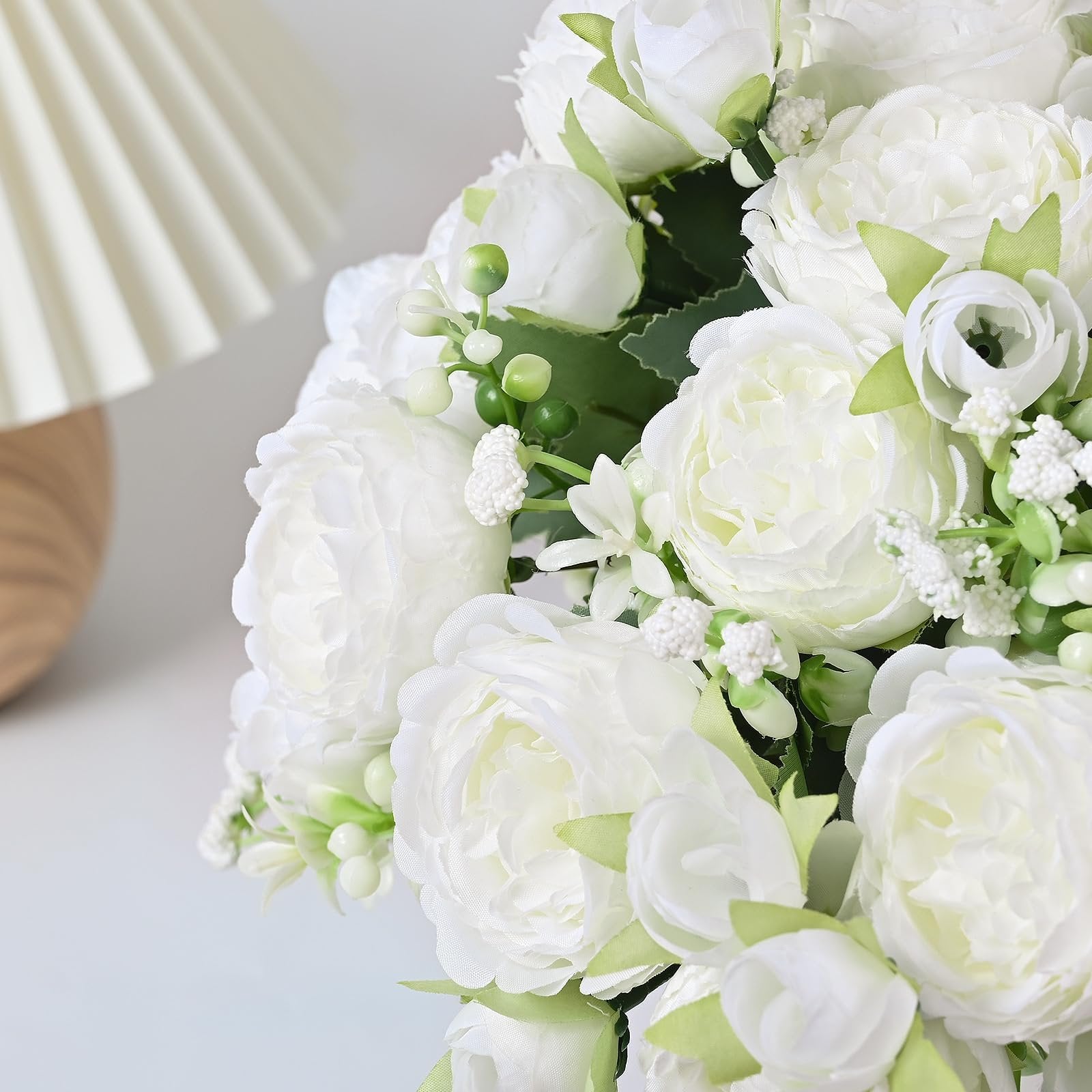 Close-up of white artificial peony bouquets with green leaves and buds, next to a pleated lamp.