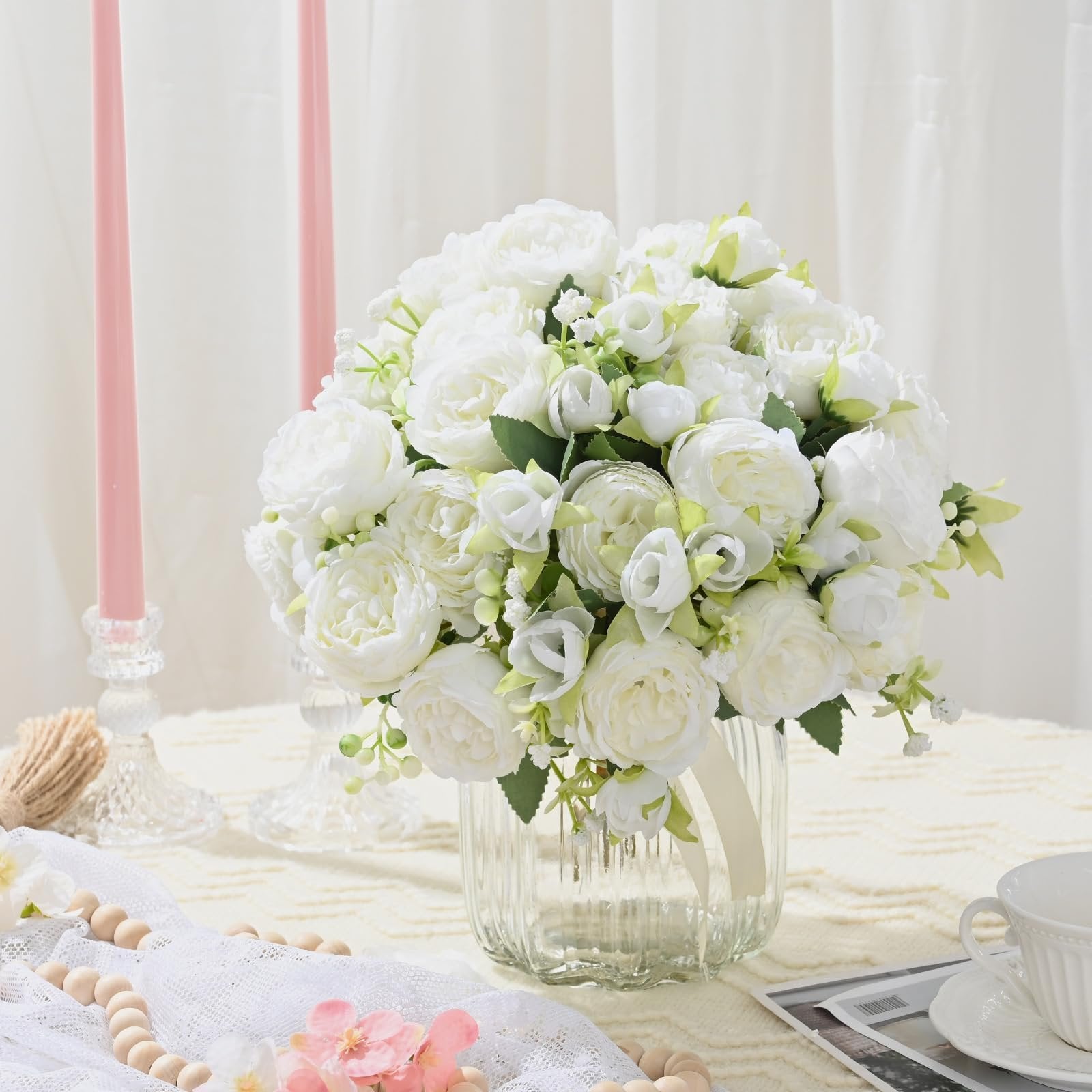 White artificial peony bouquet in a glass vase on a table with pink candles and decorative beads