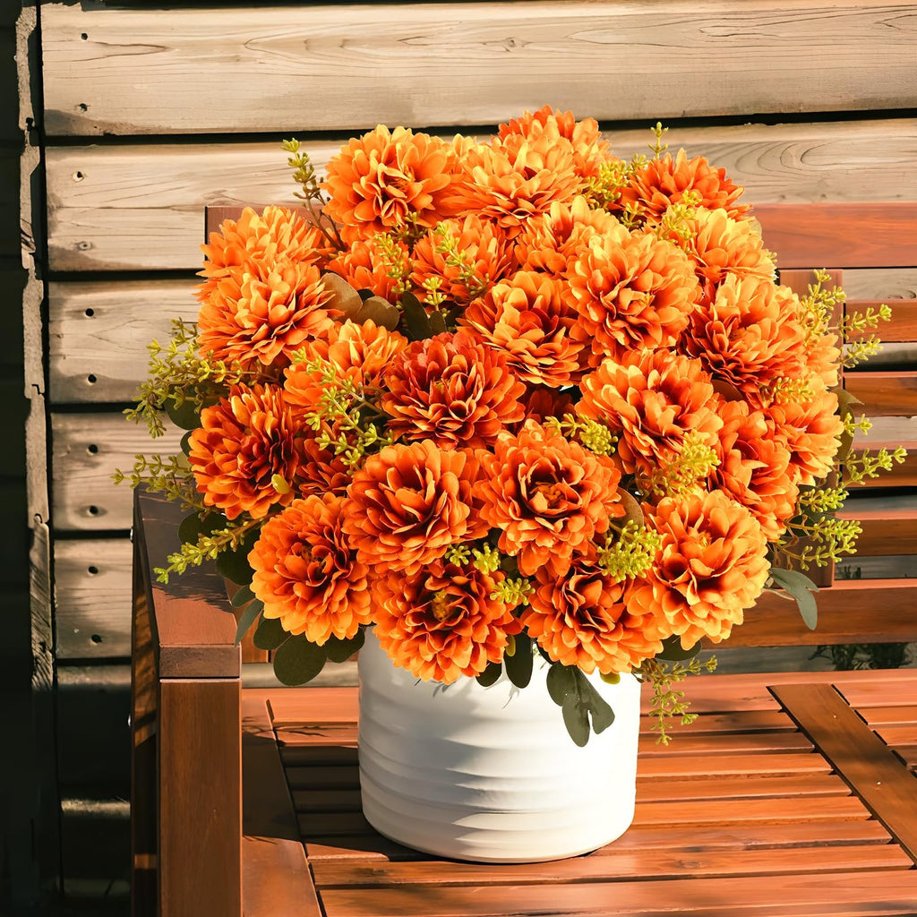 Arrangement of 36 orange artificial mums in a white vase on a wooden bench, with a wooden wall in the background.