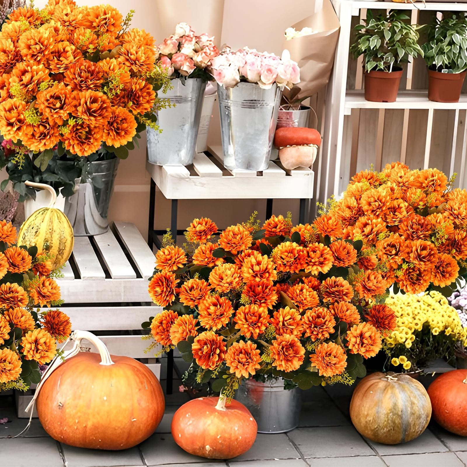 Orange artificial mums and pumpkins arranged in a fall outdoor display with potted plants in the background.