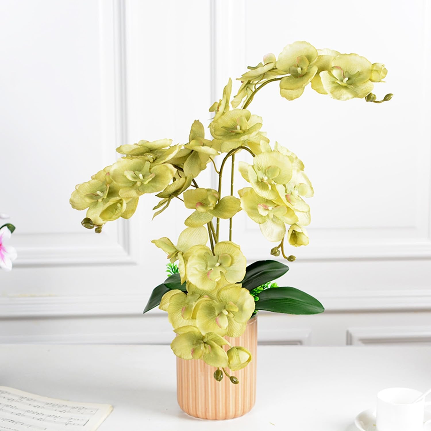 Green artificial phalaenopsis orchid stems with multiple blooms in a ribbed vase on a white surface with sheet music.