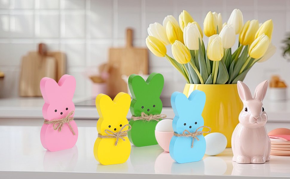 Four pastel wooden bunny figures and one pink ceramic bunny on a countertop with tulips in a yellow vase.