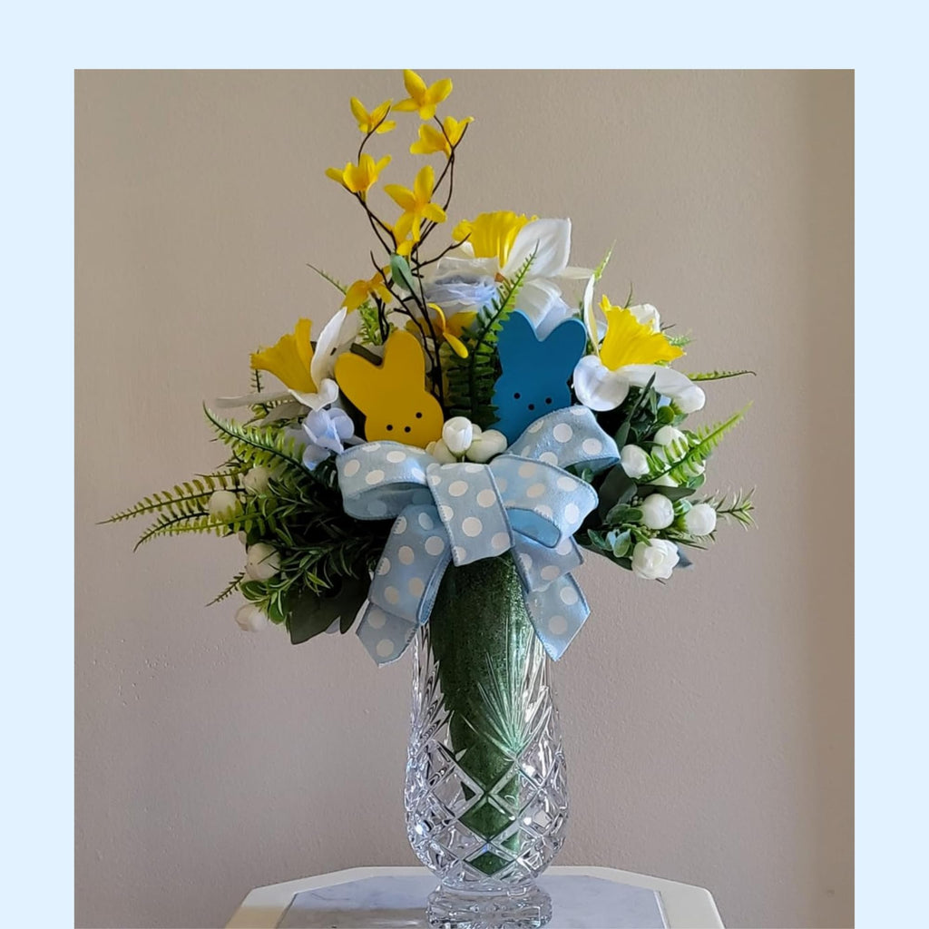 Easter floral arrangement in a crystal vase with yellow and white flowers, green foliage, and two wooden bunny figures.