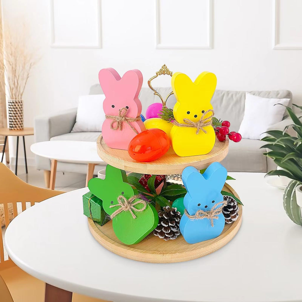 Four pastel wooden bunny decorations on a two-tiered bamboo tray, styled with Easter eggs and greenery on a table.