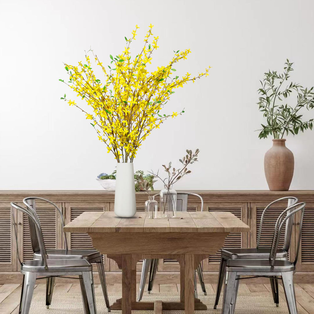 Four artificial yellow forsythia branches in a white vase on a wooden table with chairs.