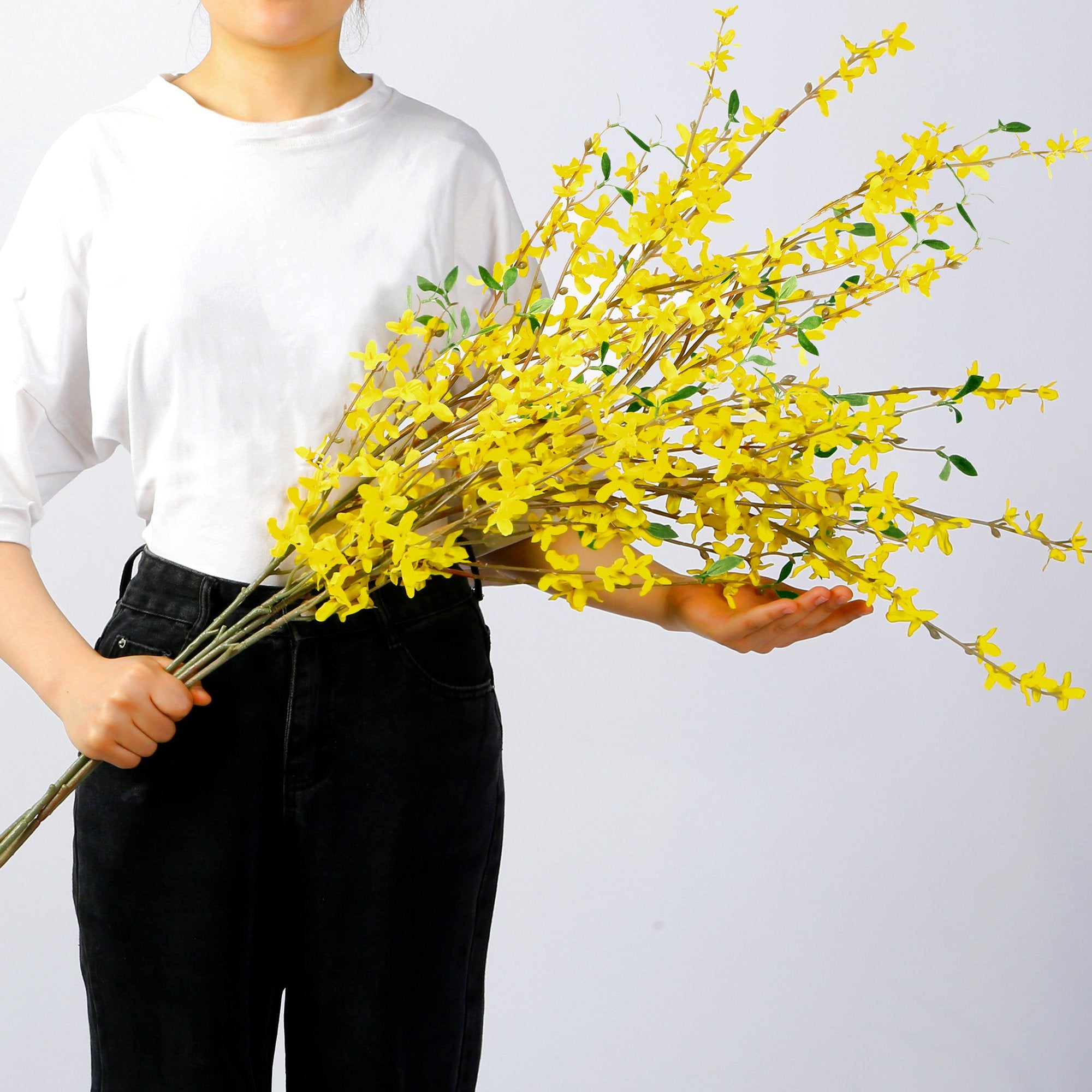 Person holding a bouquet of long, artificial yellow forsythia branches with green leaves against a white background.