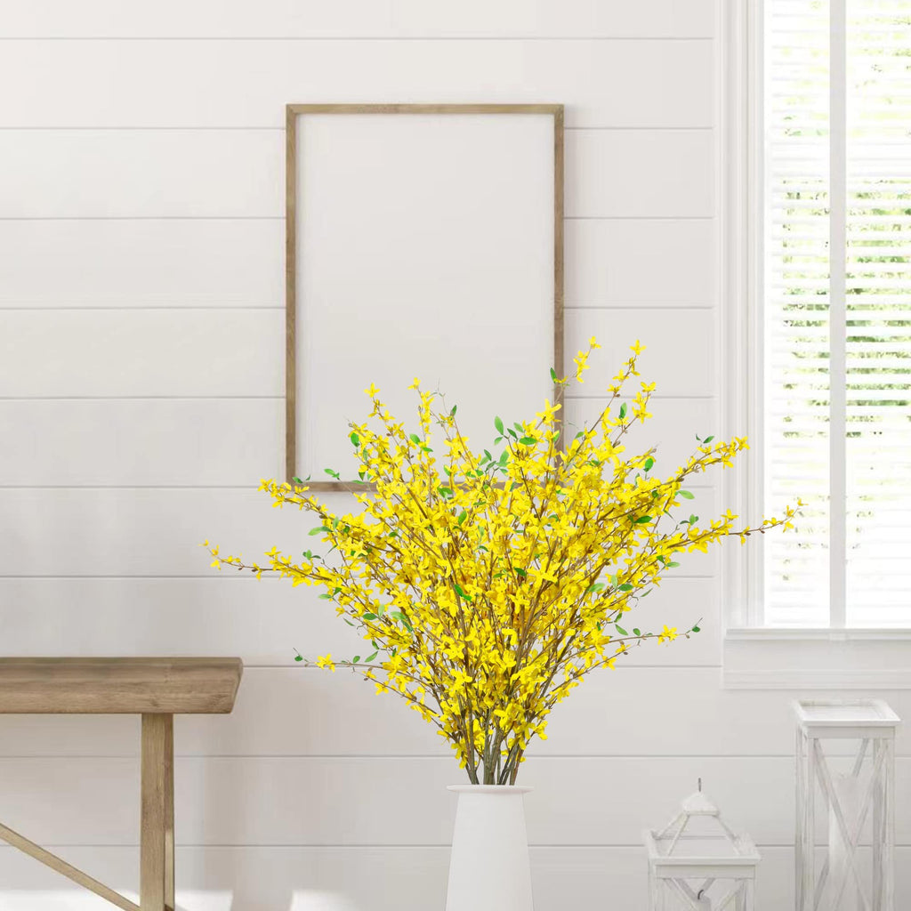 A vase of yellow silk forsythia branches with green leaves against a white shiplap wall with a framed picture and window.