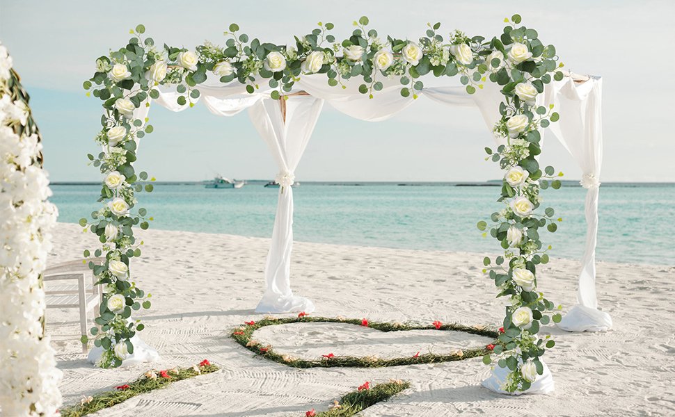 Artificial eucalyptus and white rose garland draped over a wedding arch on a beach, with a heart-shaped arrangement on the sand.
