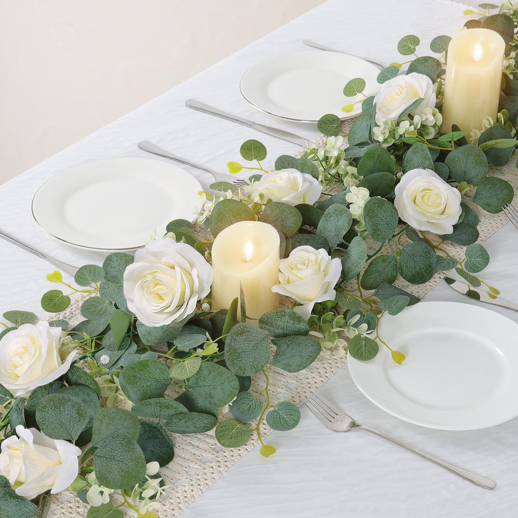 Artificial eucalyptus and white rose garland draped across a table setting with plates and candles.