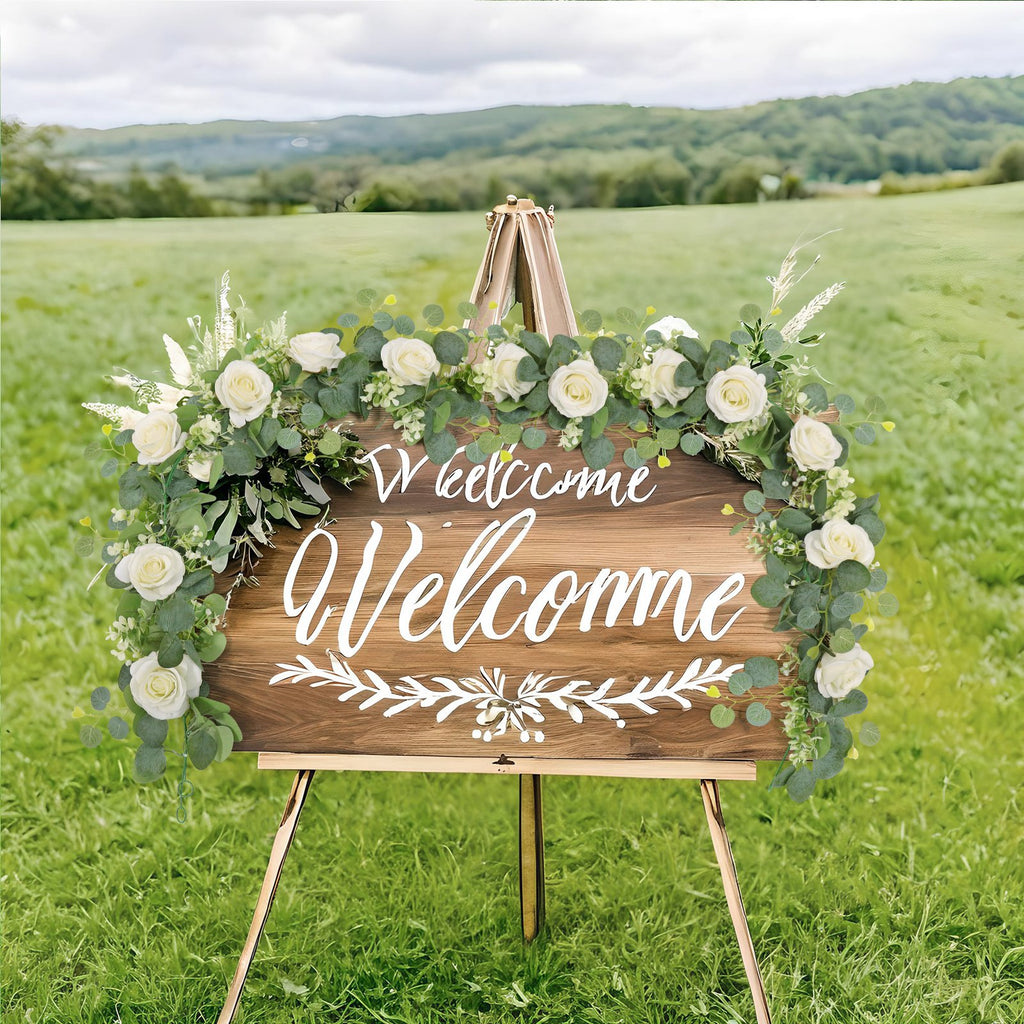 Wooden welcome sign decorated with artificial eucalyptus and white roses, displayed outdoors on an easel.