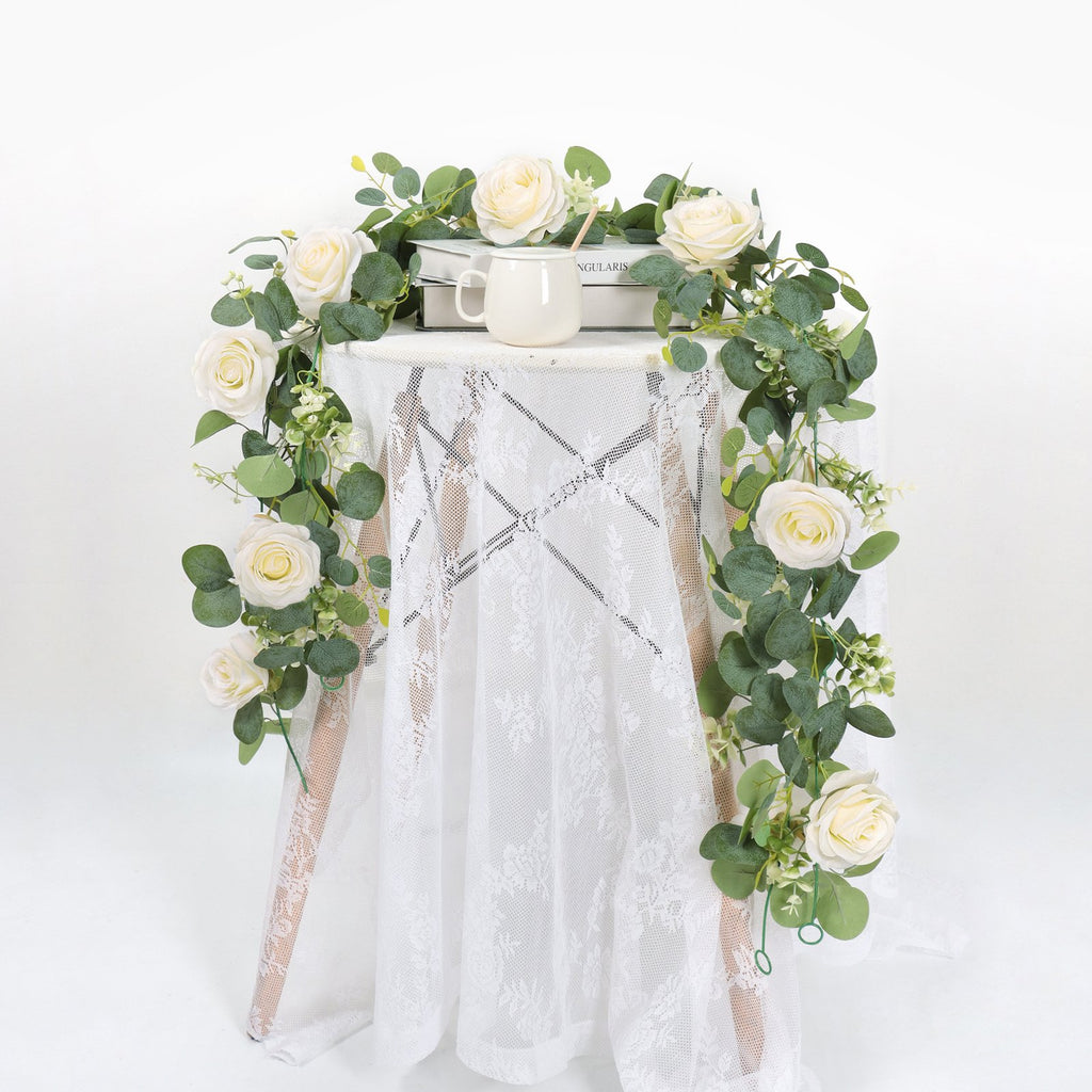 Artificial eucalyptus and white rose garland draped around a small table with books and a mug.