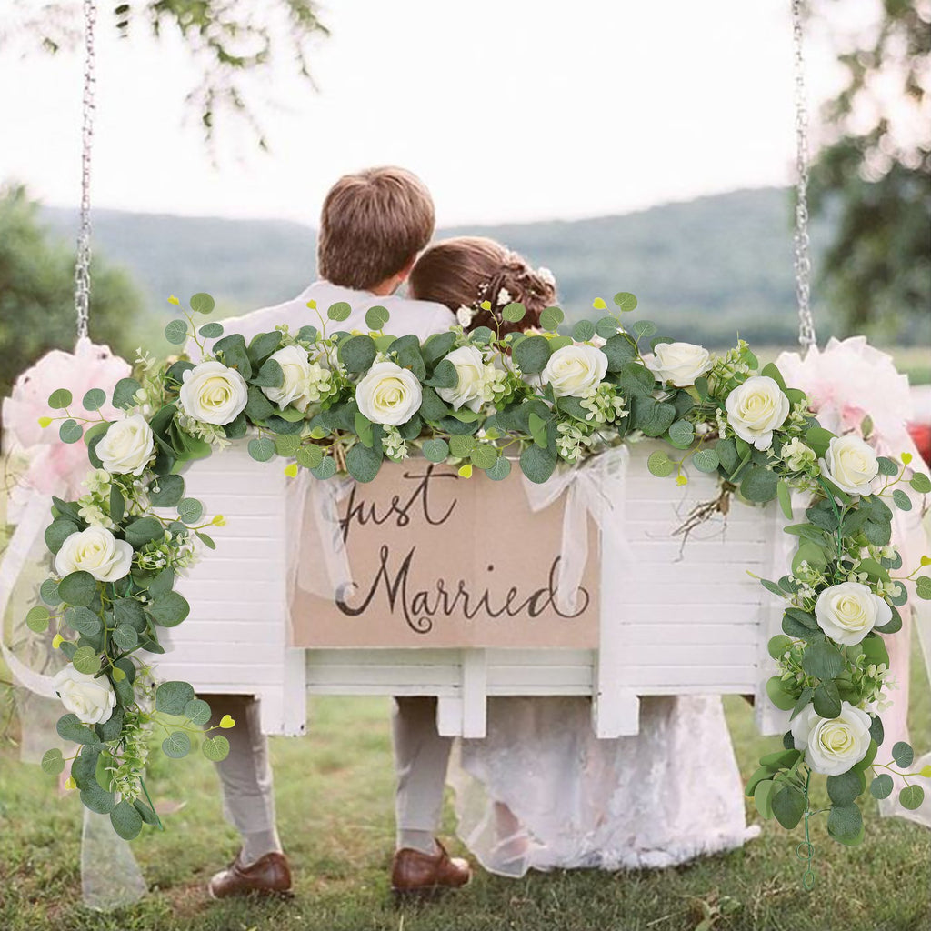 Artificial eucalyptus and white rose garland draped over a swing with a 'Just Married' sign, a couple sits on the swing.