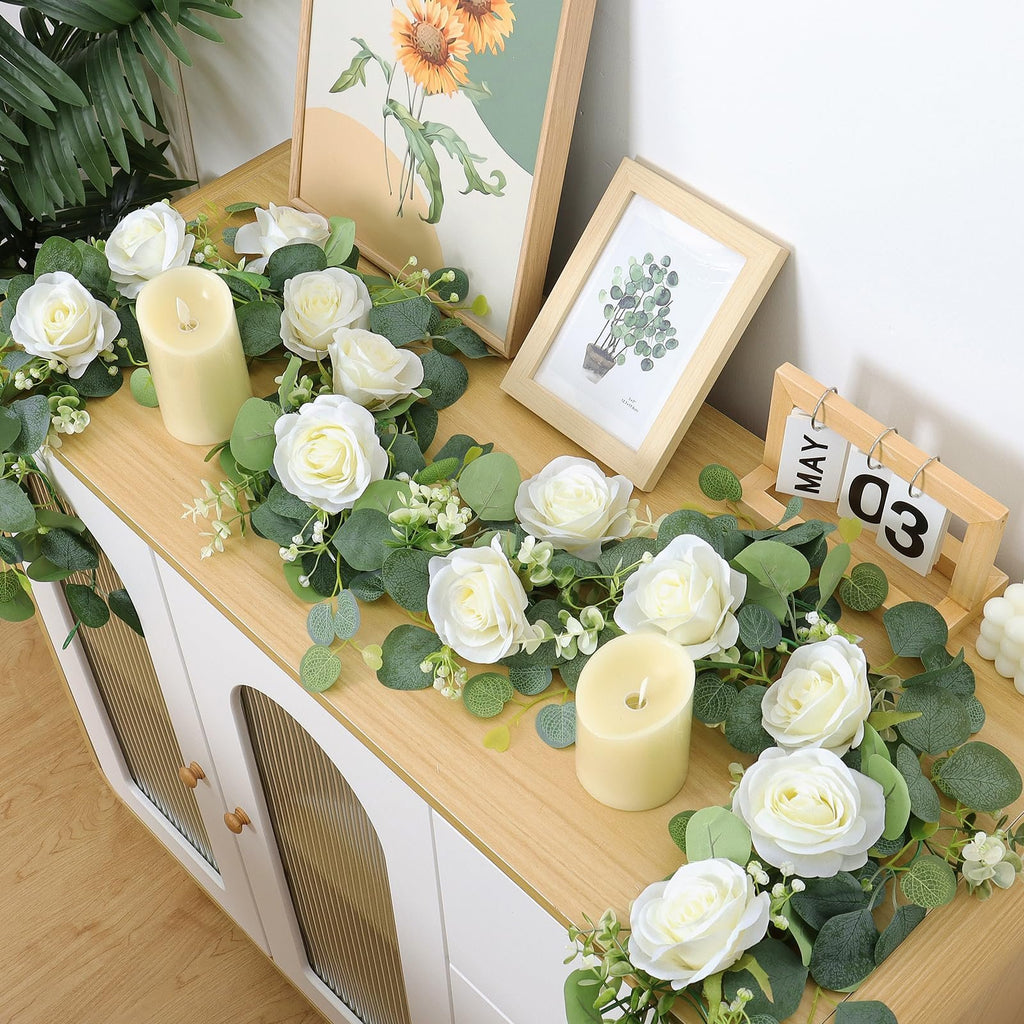 Artificial eucalyptus and white rose garland draped on a wooden surface with candles and framed art.