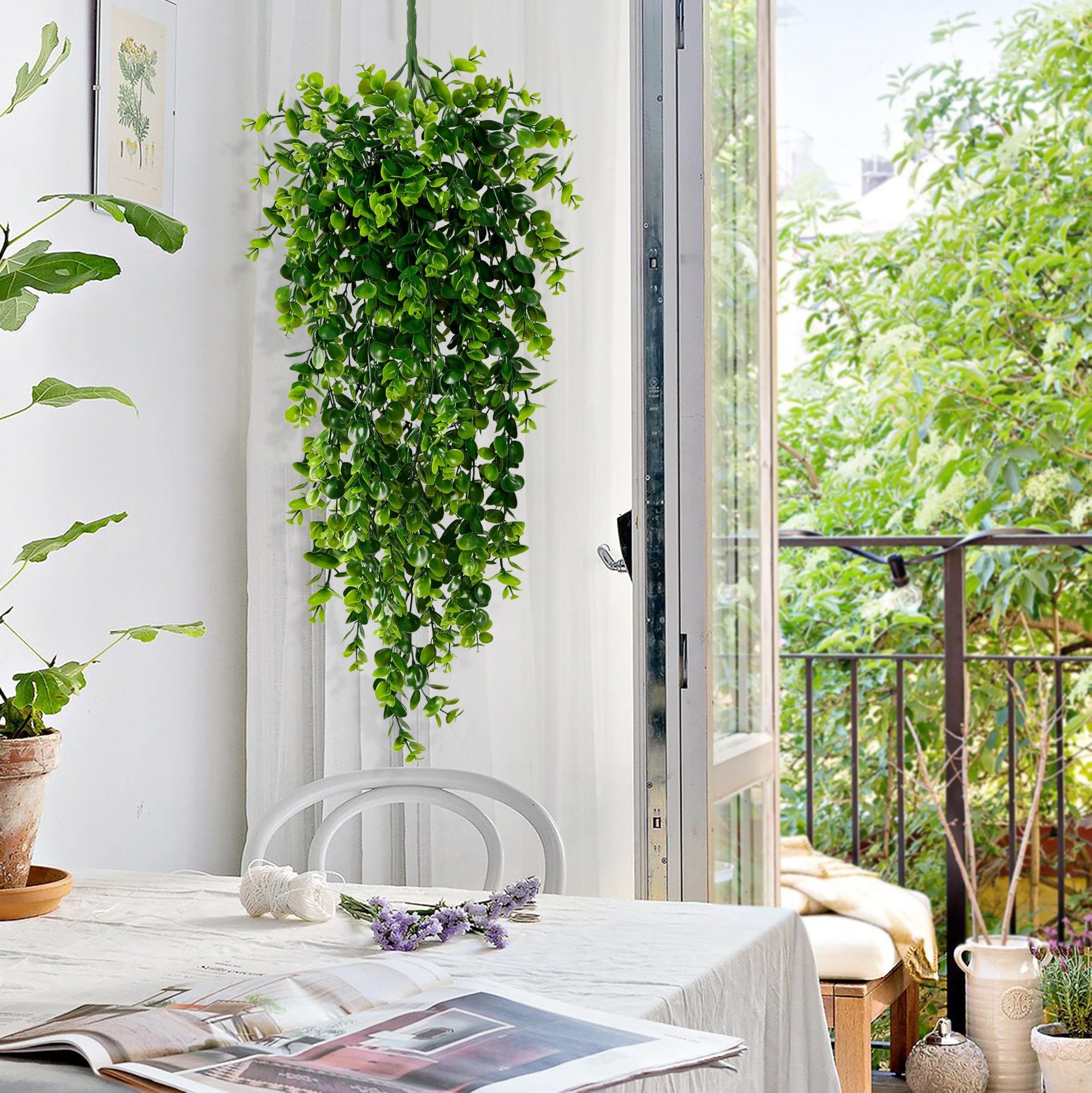 Artificial hanging eucalyptus plant with small green leaves draped over a table with a magazine and flowers