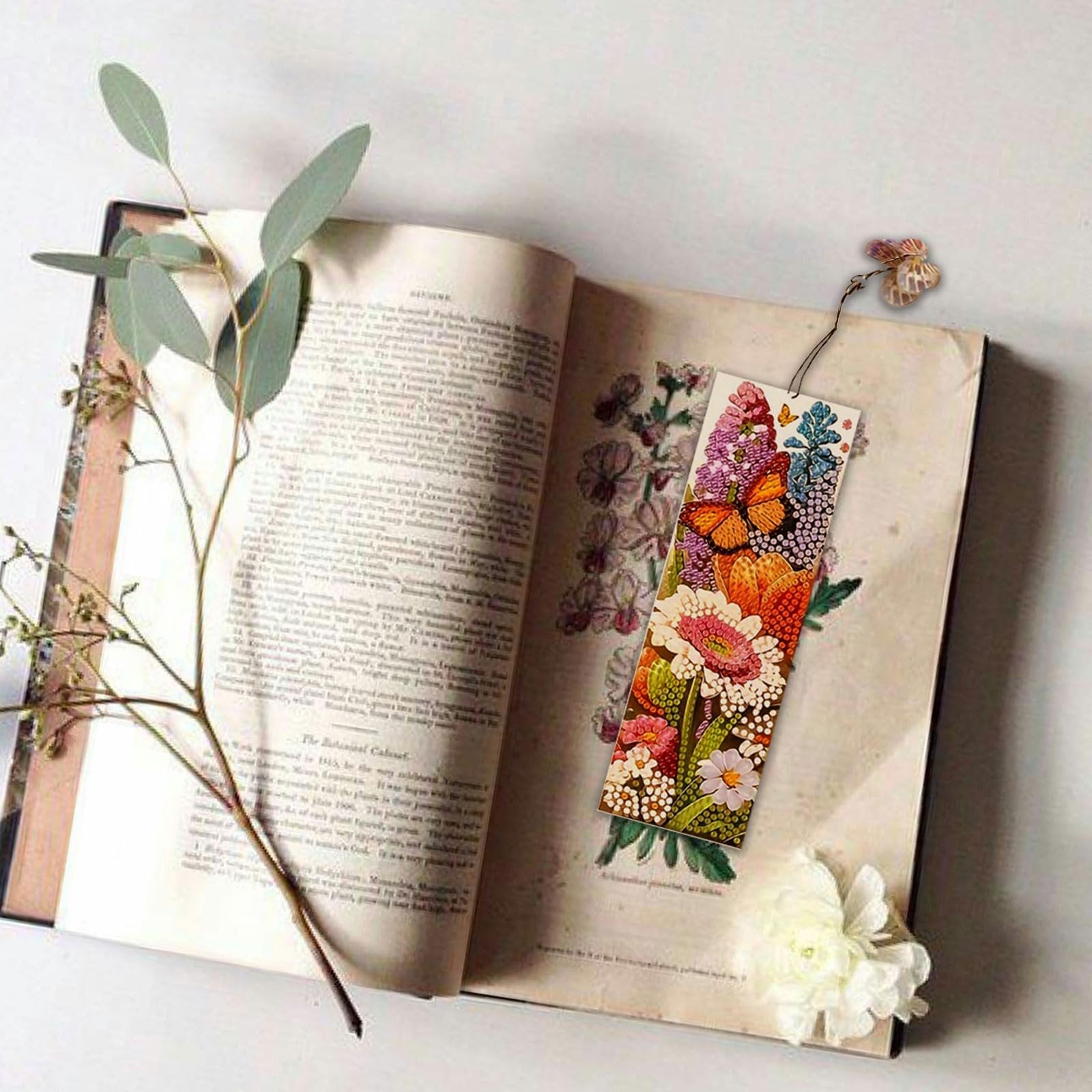 Diamond art bookmark with floral and butterfly design resting on an open vintage book with eucalyptus sprigs.