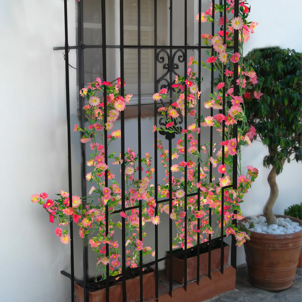 Artificial daisy flower garland with pink and rose red flowers and green leaves draped over a black metal grate in front of a window.