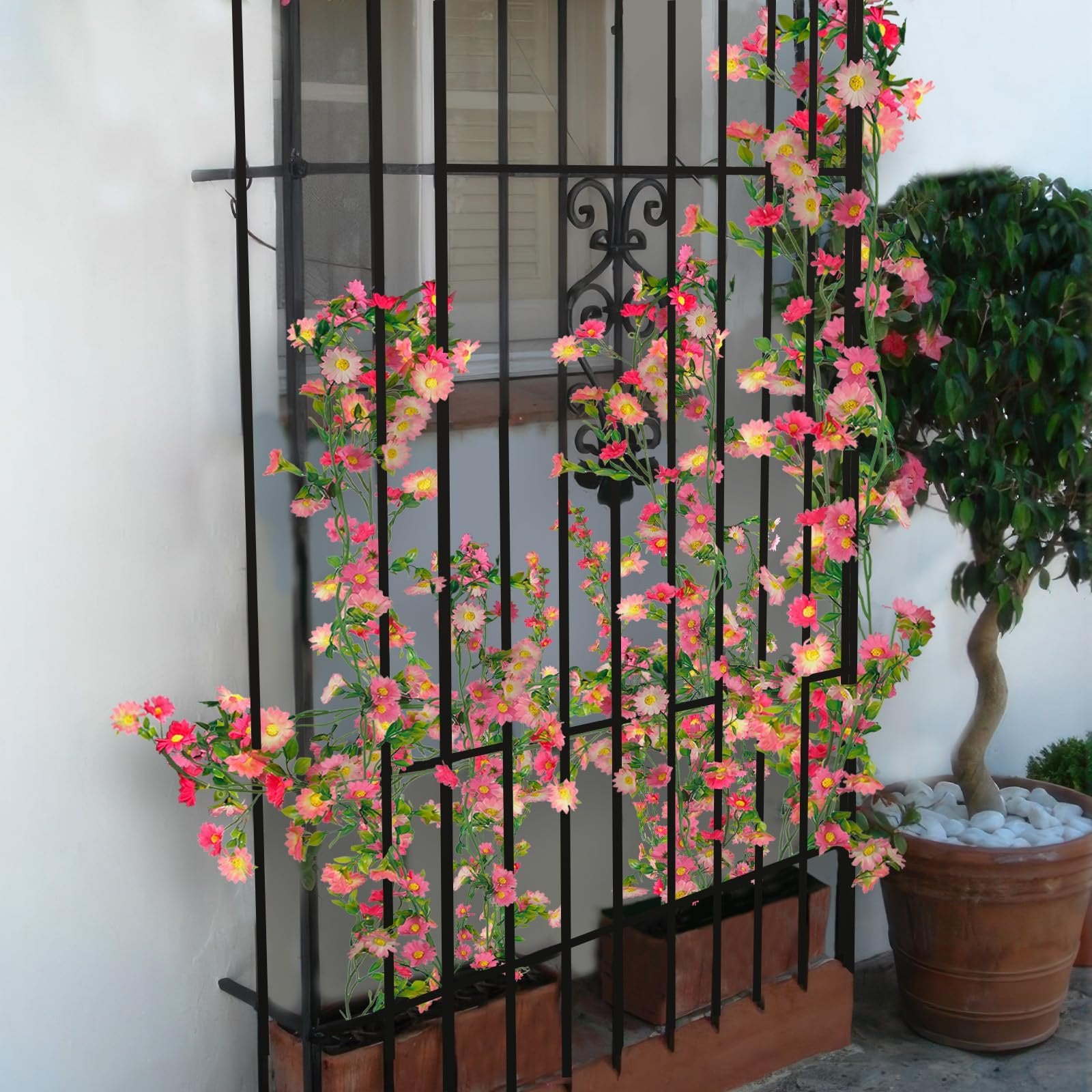 Artificial daisy flower garland with pink and rose red flowers and green leaves draped over a black metal grate in front of a window.