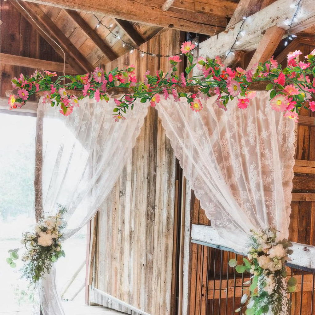 Artificial pink daisy flower garland with green leaves draped across a wooden beam with lace curtains and floral arrangements.