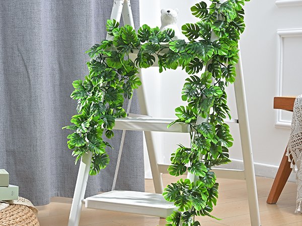 Artificial monstera leaf vines draped over a white ladder with a small white bear figurine on top.