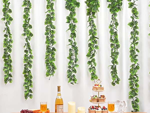 Four artificial monstera leaf vines hanging against a white backdrop, with a table set for a party below.