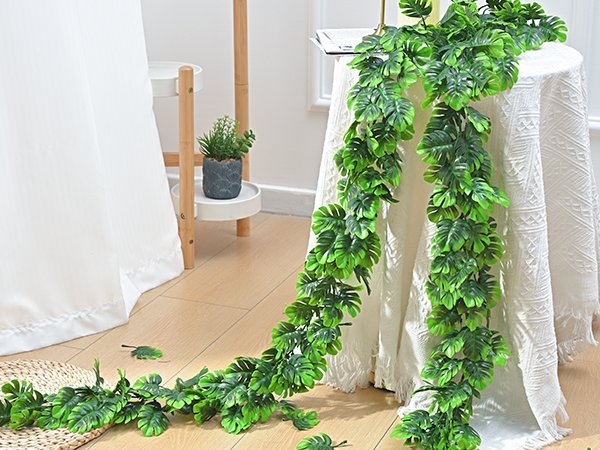 Artificial monstera leaf vines draped over a white tablecloth on a table, with a small potted plant on a shelf in the background.