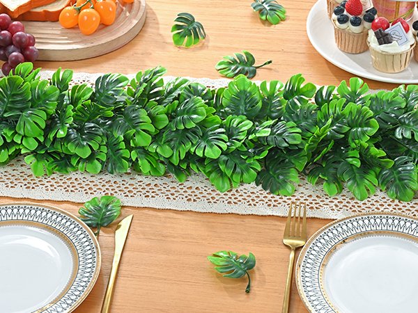 Artificial monstera leaf garland draped across a table setting with plates, cutlery, grapes, and cupcakes.