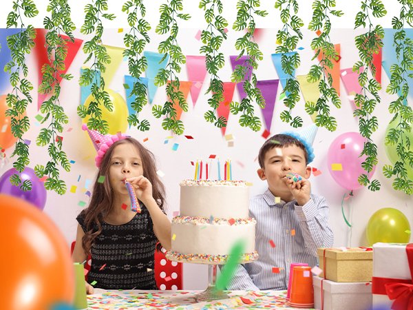 Children celebrating a birthday with a cake, balloons, and artificial monstera leaf vines hanging in the background.