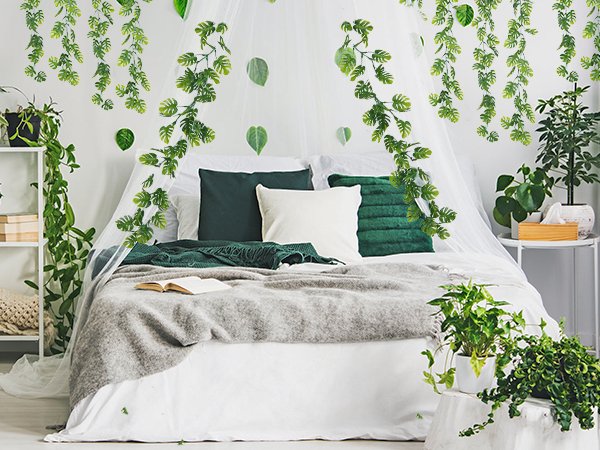 Bedroom decorated with artificial monstera leaf vines hanging from the ceiling and walls, draped over a bed with green and white pillows.