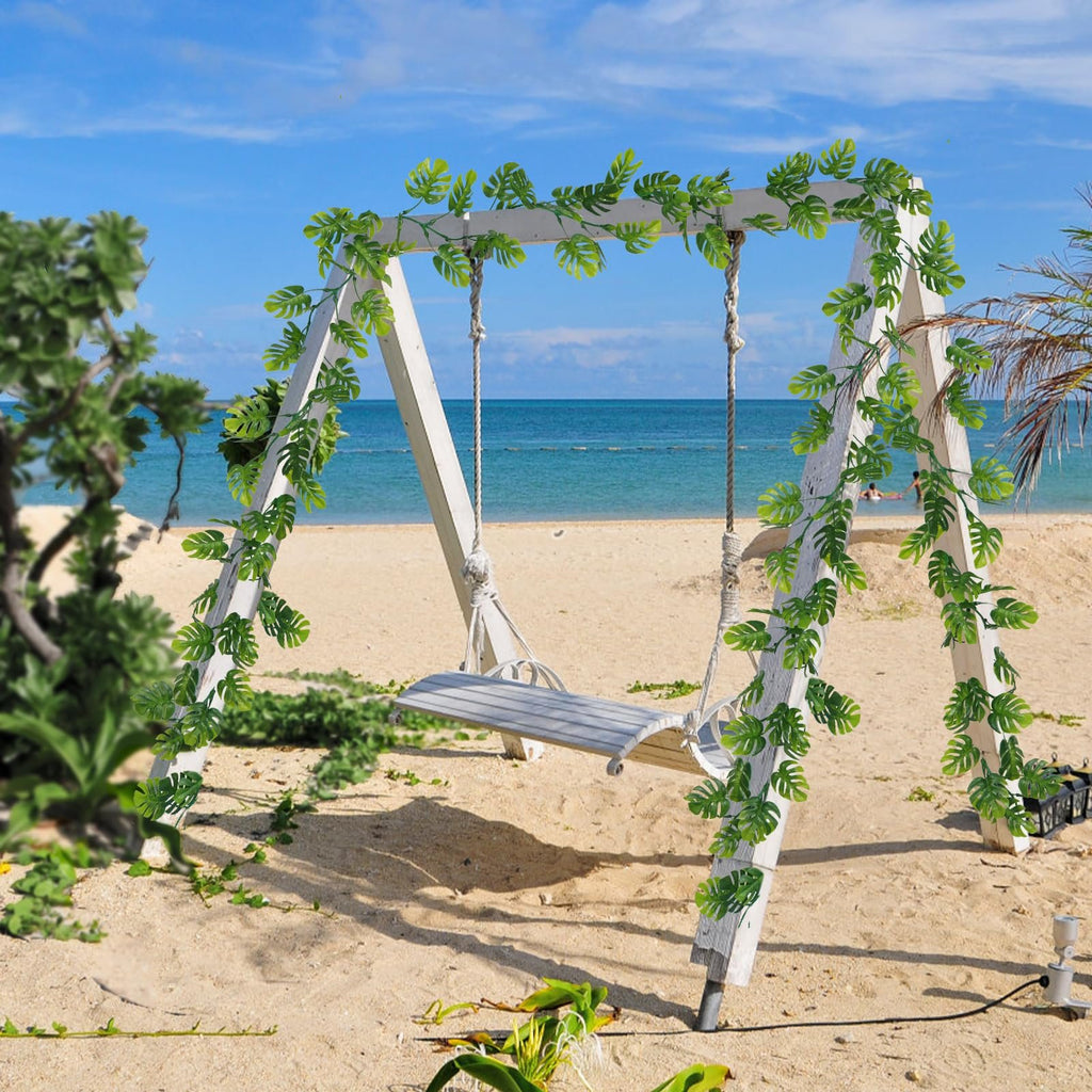 Artificial monstera leaf vines draped over a white wooden swing set on a sandy beach with the ocean in the background.
