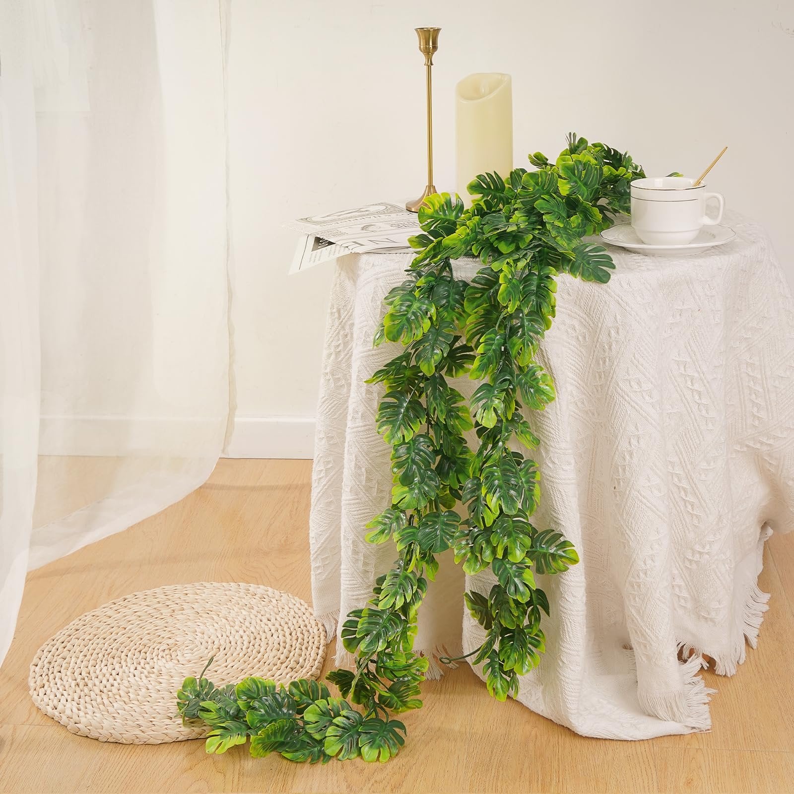 Artificial monstera leaf vines draped over a table with a cup and candlestick