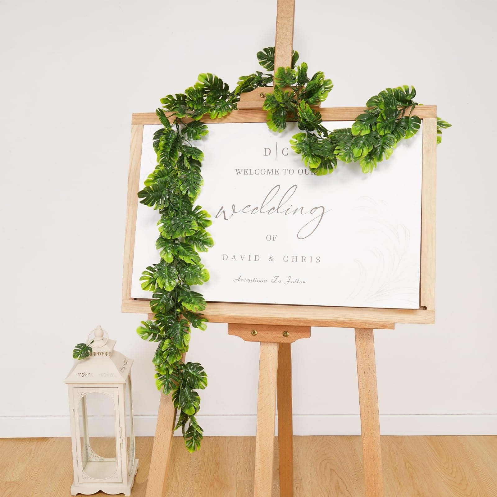 Artificial monstera leaf vine draped over a wedding welcome sign on an easel, with a decorative lantern to the side.